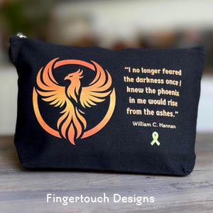 May include: Black zippered pouch featuring a phoenix design in orange and yellow within a circle. The pouch includes a quote by William C. Hannan and a green ribbon symbol. The text "Fingertouch Designs" is at the bottom.