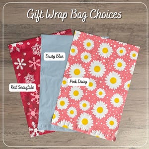 Może przedstawiać: Trzy prostokątne torebki prezentowe z napisem Gift Wrap Choices. Jedna torebka jest czerwona w białe płatki śniegu z etykietą Red Snowflake, jedna jest gładka, przygaszony błękit z etykietą Dusty Blue, a jedna jest r&oacute;żowa w biało-ż&oacute;łte stokrotki z etykietą Pink Daisy.