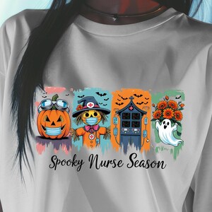 Puede incluir: Camiseta blanca con un diseño temático de Halloween. El diseño incluye una calabaza con gafas y una máscara, una enfermera espantapájaros, una casa embrujada y un fantasma con flores. El texto "Spooky Nurse Season" está impreso debajo.