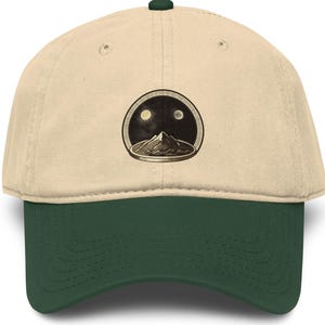 May include: A baseball cap with a cream-colored crown and a dark green brim. The cap features a circular graphic of a mountain range under two moons. The cap has a dark green button on top.