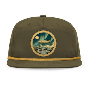 May include: An olive green baseball cap featuring a circular patch on the front. The patch displays a graphic illustration of a dirigible flying over a mountain range under a moon and aurora borealis, with a compass rose at the bottom and gold rope.