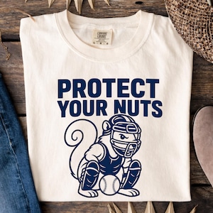 May include: Cream-colored t-shirt with the text "PROTECT YOUR NUTS" in navy blue. Below the text is a navy blue graphic of a squirrel wearing catcher's gear and holding a baseball. The shirt is folded on a wooden surface.