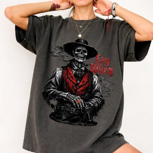 May include: Dark gray t-shirt featuring a skeletal cowboy with a hat and red vest, holding a gun. The phrase "Say When" is printed in red on the right side. The design has a vintage, distressed look.