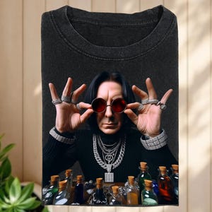 May include: Dark gray t-shirt featuring a portrait of a person wearing red-tinted sunglasses and multiple rings. The person is also wearing a black turtleneck and several necklaces. A collection of glass bottles with cork stoppers is at the bottom of the shirt.