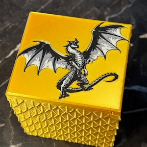 May include: A yellow box with a detailed dragon illustration on the lid. The dragon is black and white with its wings spread. The box has a textured geometric pattern on the sides.
