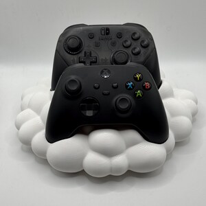 May include: Black Xbox and Nintendo Switch controllers rest on a white cloud-shaped stand. The controllers have various buttons and joysticks. The stand is a decorative piece for gaming accessories.