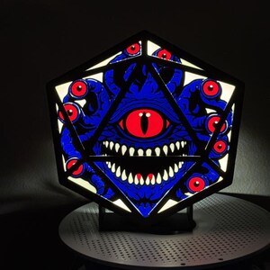 May include: A backlit, hexagonal display piece featuring a dark blue, multi-eyed creature with a large central red eye and sharp white teeth. The design is framed by a black geometric structure, creating a glowing, high-contrast visual effect.