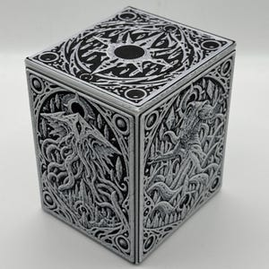 Puede incluir: Una caja rectangular con intrincadas tallas en relieve en blanco y negro en sus paneles superiores y laterales. Los diseños muestran complejos patrones orgánicos, formas geométricas nítidas y un emblema central en forma de estrella.