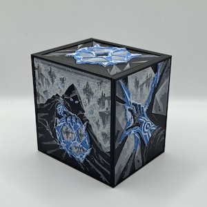 May include: A black cubic box featuring detailed illustrations on its sides and top. The artwork depicts a hooded figure and a hand reaching out, rendered in shades of black, gray, and vibrant blue, with raised, textured elements on the surfaces.