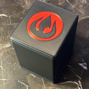 May include: A black, square box with a red emblem on the top. The emblem is a stylized flame or swirl design. The box appears to be 3D-printed and sits on a dark, marbled surface.