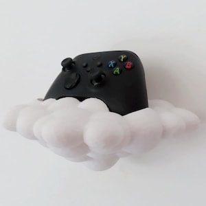 Puede incluir: Un mando de videojuegos negro descansa sobre una nube blanca y esponjosa. El mando tiene botones de colores y controles direccionales. La nube es un elemento decorativo caprichoso, que sugiere un tema lúdico.
