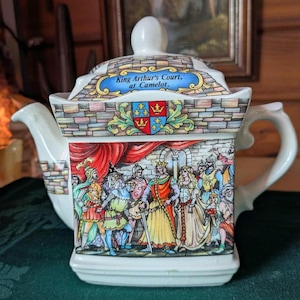 May include: A white ceramic teapot with a castle design. The lid has the text "King Arthur's Court, at Camelot." The body of the teapot shows a medieval scene with figures in period clothing. The handle and spout are curved.