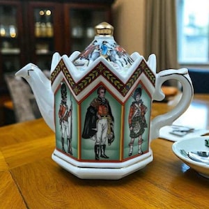 Può includere: Teiera in porcellana bianca con coperchio e manico decorativi. La teiera presenta illustrazioni di personaggi storici in uniformi militari. Il coperchio è sormontato da un pomello dorato. La teiera è appoggiata su una superficie di legno.