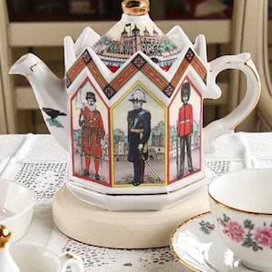 以下が含まれることがあります： 王冠型の蓋が付いた白いセラミックティーポット。歴史上の人物とロンドン塔のイラストが描かれています。ティーポットは金色のハンドルと注ぎ口があり、ティーセットの一部です。