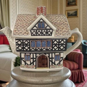Puede incluir: Una tetera decorativa de cerámica con forma de casa de estilo Tudor. La tetera presenta un tejado con diseño de ladrillos, entramado de madera en blanco y negro y detalles de ventanas azules. El asa y el pico son blancos.