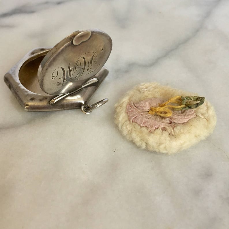 May include: A silver locket with an engraved inscription of "W.D." and a hinged lid. The locket is open and the inside is empty. A small, round, white, fluffy pad with a pink fabric centre and yellow and green fabric accents is next to the locket.