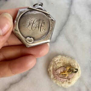 May include: A silver locket with a hexagonal shape and a hinged lid. The locket has an engraved heart and the initials "HCM" on the front. The locket is open and a small, round, white and pink fabric pad is visible.