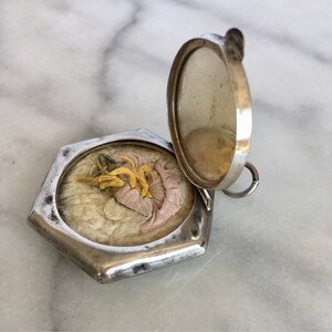 May include: A silver locket with a hexagonal shape and a hinged lid. The locket is open, revealing a small, white, fluffy pad with a pink and yellow fabric flower on top.