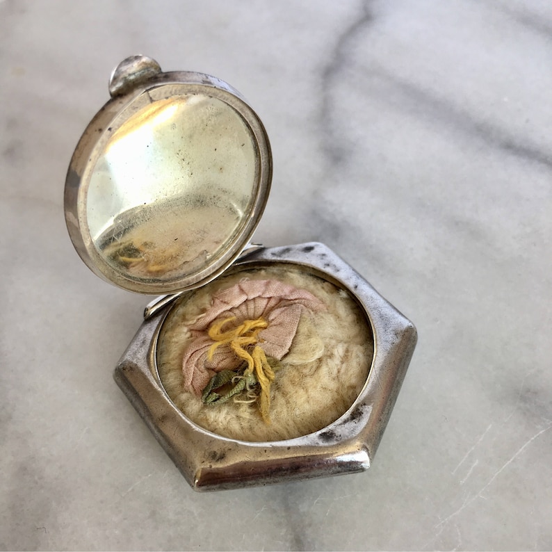 May include: A silver compact with a hinged mirror and a floral design on the inside. The compact is open and the floral design is visible.