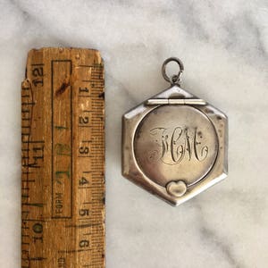 May include: A silver hexagonal locket with a hinged lid and a heart-shaped charm. The locket is engraved with the initials "J.C.M."