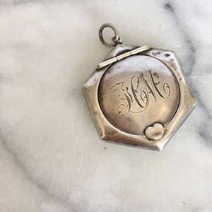 May include: A silver locket with a hexagonal shape and a hinged lid. The locket has an engraved heart and the initials "J.W.M." on the front.