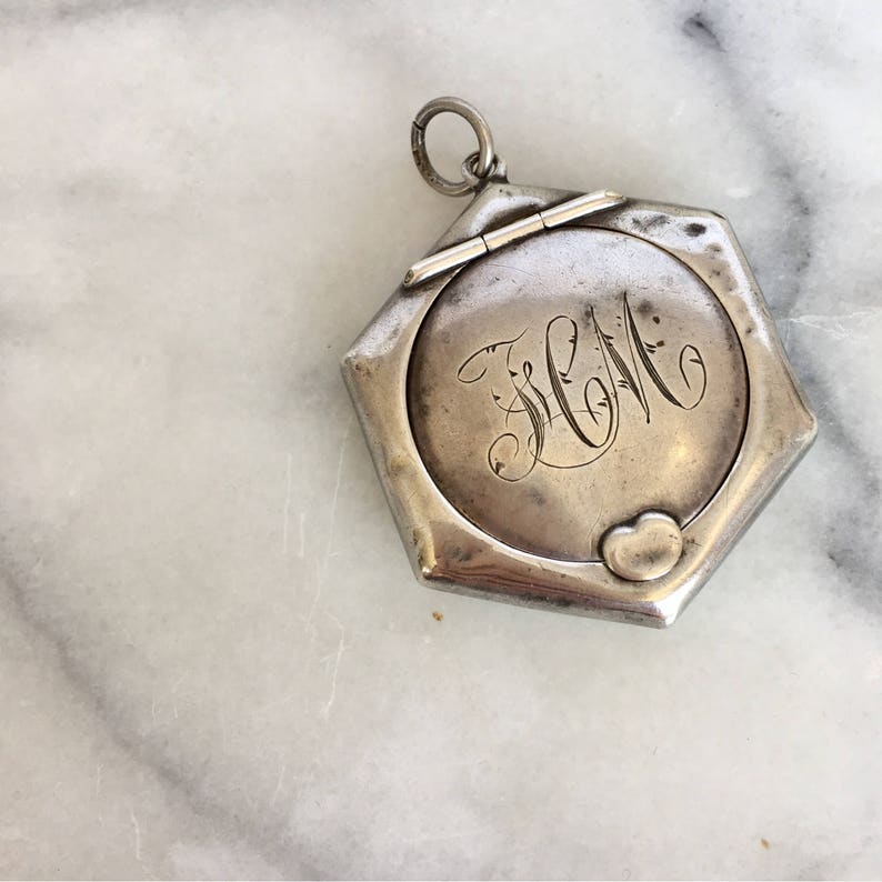 May include: A silver locket with a hexagonal shape and a hinged lid. The locket has an engraved heart and the initials "J.W.M." on the front.