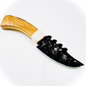 May include: A knife featuring a black, serrated obsidian blade with a glossy finish. The handle is made of light-coloured, polished wood with a visible grain, connected to the blade by a white, tightly wrapped cord binding at the base of the handle.