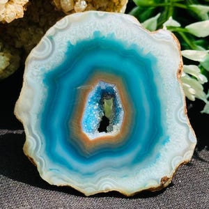May include: A polished agate slice, showcasing concentric bands of white, teal, and deep blue. The centre has a crystalline cavity with orange and brown hues. The natural stone is set against a dark backdrop, accentuating its vibrant colours.