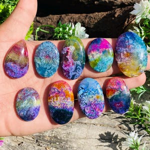 May include: Nine polished gemstone cabochons in oval and teardrop shapes. The stones display blues, purples, oranges, and greens, with intricate patterns. The gemstones are held in a hand. The stones are likely to be measured in centimetres.