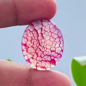 Puede incluir: Una gema ovalada con un patrón agrietado en forma de telaraña en tonos rosa y blanco. La piedra se sostiene entre dos dedos sobre un fondo azul claro. El diseño único de la gema es el punto focal.