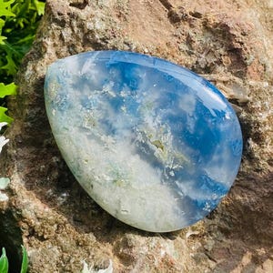 Könnte beinhalten: Ein polierter, oval geformter Edelstein mit einem auffälligen blau-weißen Muster, das an Wolken am Himmel erinnert. Der Stein hat eine glatte, glänzende Oberfläche und liegt auf einem rauen, braunen Felsenhintergrund. Ein natürlicher Dekorationsartikel.