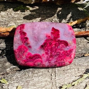 May include: A rectangular gemstone with rounded edges. The stone displays a mix of pink and magenta shades, with a swirling pattern of deep magenta. The stone is set against a natural wood background, with white flowers and greenery in the background.