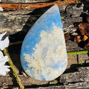 May include: A teardrop-shaped gemstone with a blue and white cloud-like pattern. The polished stone has a smooth surface and is set against a rustic wooden background. The stone's colors and patterns evoke a sense of the sky.