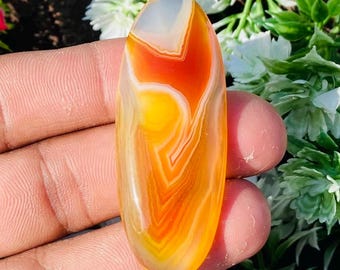 May include: An elongated, polished gemstone with a vibrant color gradient. The stone transitions from a translucent white at the top to a warm orange and yellow, showcasing natural banding patterns. The gemstone is held in a hand, with a blurred background of greenery.