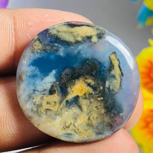 Könnte beinhalten: Ein runder, polierter Edelstein-Cabochon mit einer durchscheinenden, himmelblauen Grundfarbe. Der Stein zeigt komplexe, moosartige Einschlüsse in Senfgelb, Anthrazit und Schwarz, die ein natürliches, landschaftsähnliches Muster bilden.