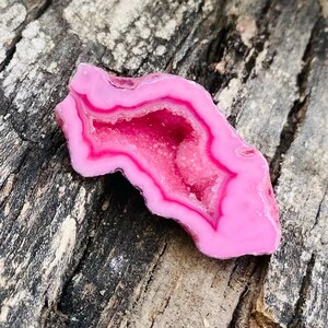 以下が含まれることがあります： 鮮やかなピンクのアゲートジオードのスライスで、結晶質の中心部があります。ジオードは明るいピンク色の外縁と、より明るいピンク色の内部を持ち、自然な模様と質感を表現しています。風化した木製の表面に石が展示されています。