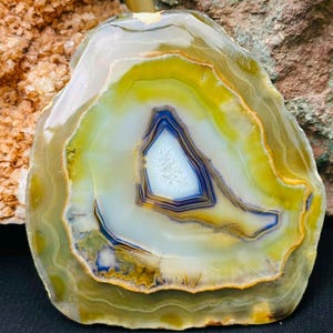 May include: A polished agate slice featuring concentric bands of translucent yellow, olive green, and white, surrounding a central crystalline cavity with deep blue and white mineral deposits, showcasing natural geological patterns and smooth edges.