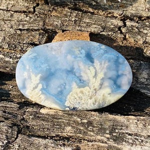 May include: An oval-shaped gemstone with a cloudy blue and cream pattern, resembling a landscape. The stone is set against a textured, brown wooden surface, with white flowers in the background.