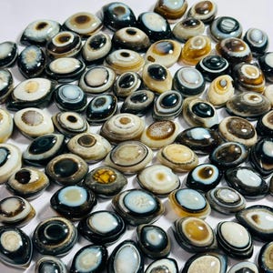 May include: A collection of numerous oval-shaped agate stones, each featuring distinct concentric bands of colour including white, black, brown, and tan. These polished cabochons display natural eye-like patterns with smooth, rounded surfaces.