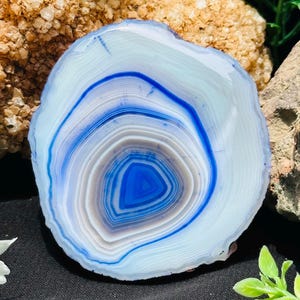 Op de afbeelding: Een cirkelvormige schijf van agaatsteen met concentrische banden in levendig blauw, wit en doorschijnend grijs. Het gepolijste oppervlak toont een duidelijk gelaagd patroon dat uitstraalt vanuit een diepblauw centrum, met natuurlijke mineraalformaties.