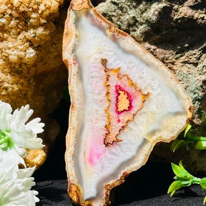 Op de afbeelding: Een driehoekige agaatplak met een doorschijnend wit kristallijn centrum en een levendige roze en oranje gekartelde kern. De steen heeft een ruwe, aardebruine buitenrand en vertoont natuurlijke minerale banden over het oppervlak.