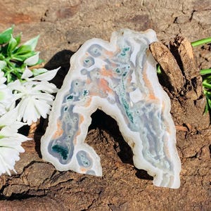 May include: A polished agate stone with an irregular shape, displaying natural patterns. The stone features bands of white, grey, orange, and green. It is presented on a textured, brown wooden surface, highlighting its unique colours and organic form.