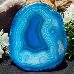 Può includere: Una fetta di agata blu lucida e traslucida caratterizzata da bande concentriche di varie tonalità di blu e bianco. La pietra presenta un bordo esterno naturale frastagliato e una superficie piatta e liscia con complessi motivi minerali.