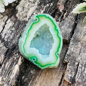 May include: A polished, geode stone with a vibrant green and white border, surrounding a crystal-filled interior with shades of blue and white. The stone is set against a weathered, wooden surface, with white flowers in the background.