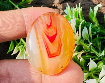 May include: Oval-shaped polished agate gemstone. The stone features layers of orange, red, and white hues. The gemstone is held between fingers, with greenery and a wood-like background.