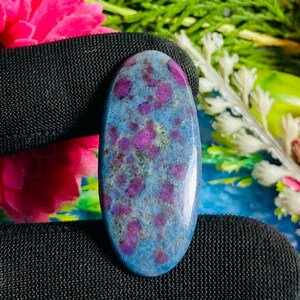 Op de afbeelding: Een ovaalvormige edelsteen met een blauwe basis en levendige roze insluitsels. De gepolijste steen vertoont een gespikkeld patroon, met de roze elementen verspreid over de blauwe matrix. De edelsteen wordt tegen een zwarte achtergrond gehouden, met bloemelementen op de achtergrond.