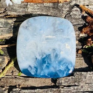Può includere: Una gemma quadrata e lucidata con un gradiente di tonalità blu e bianche, che ricorda le onde dell'oceano. La pietra è posta su una superficie di legno marrone strutturata, con fiori bianchi sullo sfondo.