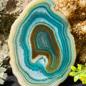 Op de afbeelding: Een gepolijste agaatplak met concentrische banden in blauwgroen, turkoois en donkerbruin. De steen heeft een doorschijnende buitenrand met een gladde, ronde vorm en toont natuurlijke minerale lagen die variëren van donkerbruin tot blauw.