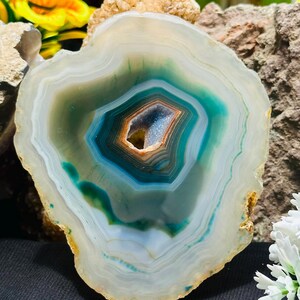 Op de afbeelding: Een gepolijste agaatplak met concentrische banden van wit, doorschijnend grijs en levendig groenblauw. Het midden bevat een kleine, holle geode-holte bekleed met kleine, sprankelende blauwe kristallen, omringd door een dunne, warmbruine ring.
