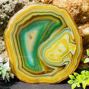 May include: A circular agate slice featuring concentric bands of vibrant yellow, deep emerald green, and soft teal. The centre displays a small, crystalline geode cavity with sparkling mineral deposits, surrounded by intricate, layered natural patterns.